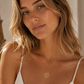 A woman with blonde hair wearing a delicate gold necklace with a sunburst pendant, styled with a white spaghetti strap top.