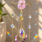 A gold-toned metal suncatcher wind spinner hanging in a window, featuring a pink crystal, a yellow crystal, and multiple iridescent clear crystals that catch and refract sunlight into rainbow patterns on the wall and tabletop.