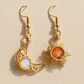 A pair of gold wire-wrapped dangle earrings on a textured beige background. One earring features a crescent moon shape wrapping around a round iridescent white-blue stone, and the other features a sunburst design wrapping around a round orange stone.