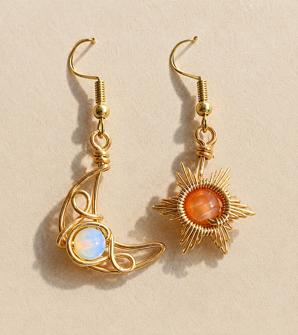 A pair of gold wire-wrapped dangle earrings on a textured beige background. One earring features a crescent moon shape wrapping around a round iridescent white-blue stone, and the other features a sunburst design wrapping around a round orange stone.
