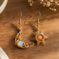 A pair of gold-toned dangle earrings displayed on a wooden surface. One earring features a crescent moon shape with a light blue opal-like stone, and the other features a sunburst design with an orange stone.