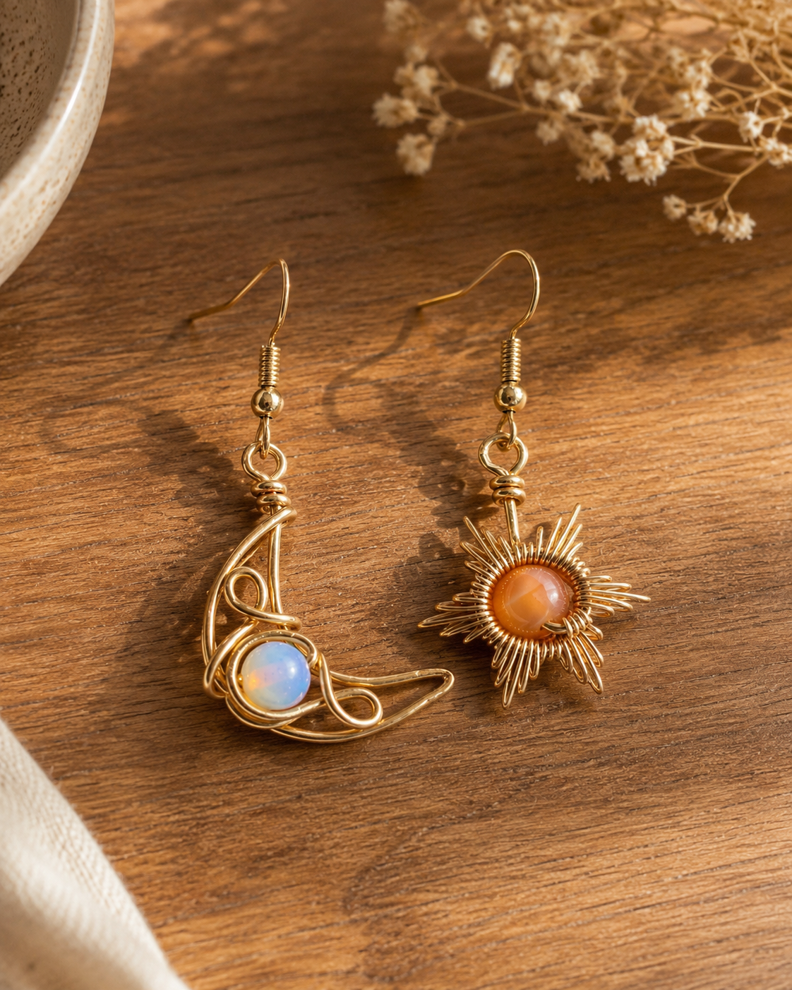 A pair of gold-toned dangle earrings displayed on a wooden surface. One earring features a crescent moon shape with a light blue opal-like stone, and the other features a sunburst design with an orange stone.