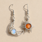 A pair of handmade silver dangle earrings on a beige background. One earring features a polished silver crescent moon wrapping around a round iridescent blue-white opal.