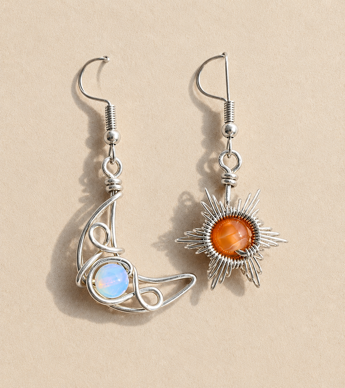 A pair of handmade silver dangle earrings on a beige background. One earring features a polished silver crescent moon wrapping around a round iridescent blue-white opal.