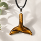 A tiger's eye pendant shaped like a whale tail, suspended from a black braided cord with a silver-toned metal cap, displayed on a light beige textured surface with soft leaf shadows.