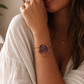 A close-up lifestyle shot of a woman wearing a gold-toned double-band cuff bracelet with a central purple amethyst crystal tree motif.